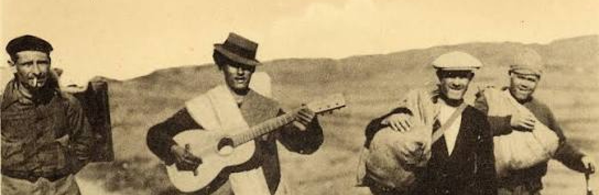 4 hommes marchent. L'un porte une guitare, les 3 autres des sacs. C'est une photographie assez ancienne des années 40.