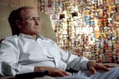 Un homme est assis sur un fauteuil et un mur rempli de photos est derrière lui. 