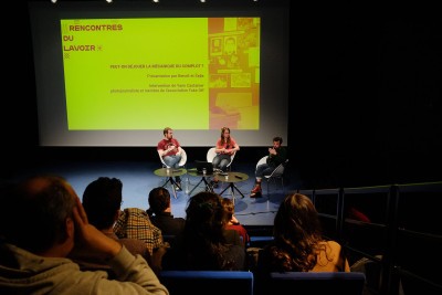 En configuration conférence, 3 personnes sur un plateau assises sur des fauteuils, le public assis dans des gradins est vu de dos. Sur l'écran : "Rencontres du lavoir : peut-on déjouer la mécanique du complot ?" - Agrandir l'image 2 sur 6, fenêtre modale