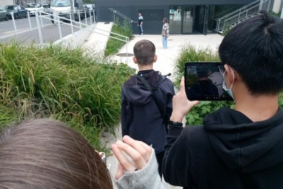 Des enfants vus de dos filment d'autres enfants au loin devant le parvis d'un bâtiment - Agrandir l'image 3 sur 6, fenêtre modale